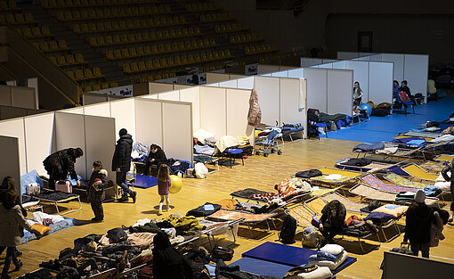 In einer Turnhalle wurden Liegen aufgestellt, damit die Menschen nach ihrer langen Flucht ausruhen können. In einer Turnhalle wurden Liegen aufgestellt und durch Trennwände separiert, damit Menschen sich ausruhen können.