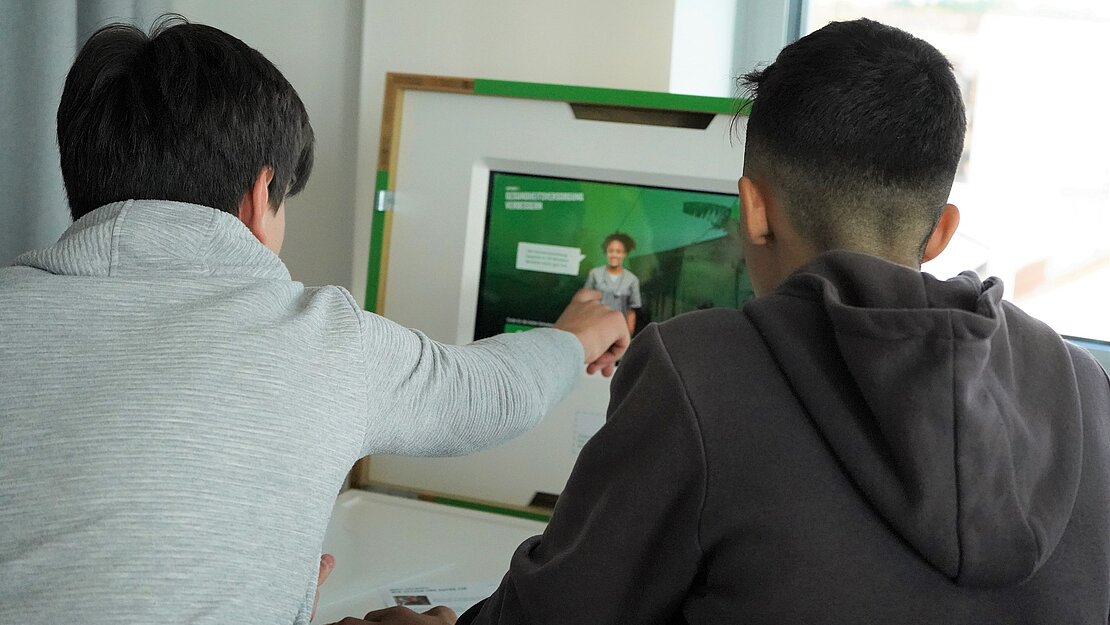 Zwei Jungen sitzen vor einer aufgeklappten Box mit Bildschirm. Einer der Jungen drückt auf den Touchscreen.