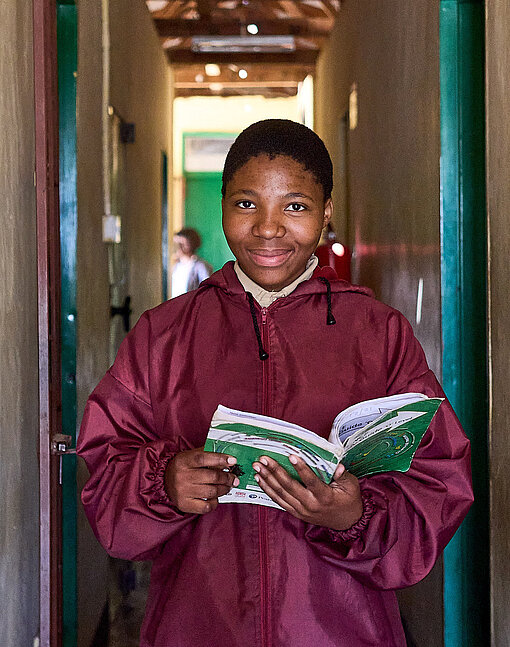 Mädchen in roter Jacke steht im Flur und hat ein aufgeschlagenes Buch in der Hand