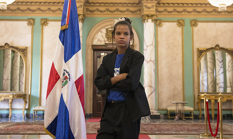 Ein Mädchen steht mit verschränkten Armen in einem Saal, neben ihm die dominikanische Flagge