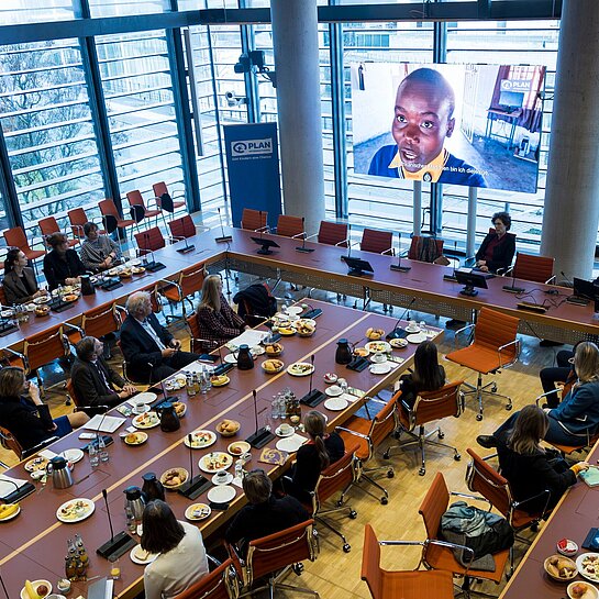 Foto von oben mit Blick auf die Tische, an denen die Teilnehmenden sitzen, und einen großen Bildschirm, auf dem ein Film mit Mädchen aus Simbabwe abgespielt wird