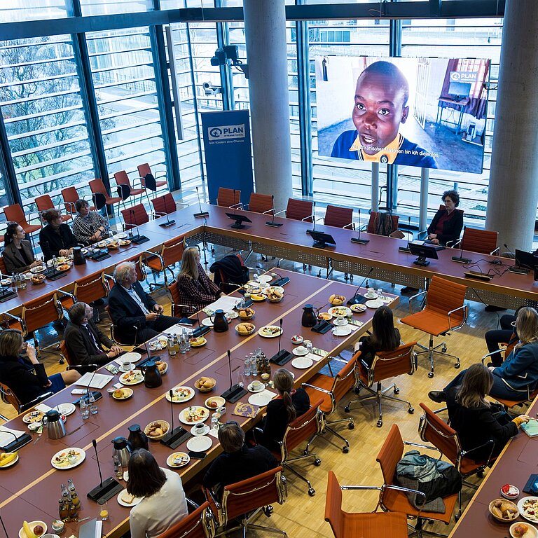 Foto von oben mit Blick auf die Tische, an denen die Teilnehmenden sitzen, und einen großen Bildschirm, auf dem ein Film mit Mädchen aus Simbabwe abgespielt wird