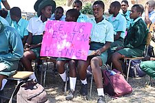 Schülerinnen sitzen in Stuhlreihen und eine von ihnen hält ein Schild auf dem "Give us education not husbands" steht, in den Händen.