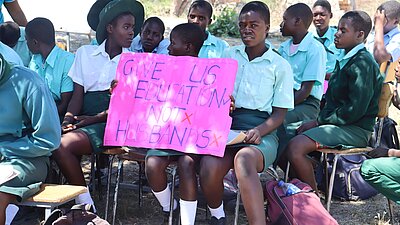 Schülerinnen sitzen in Stuhlreihen und eine von ihnen hält ein Schild auf dem "Give us education not husbands" steht, in den Händen.