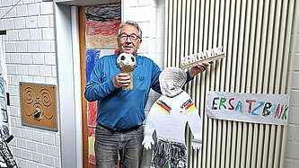 Ein Mann in blauem T-Shirt hält einen World-Cup-Pokal aus Pappe in die Kamera und lacht.