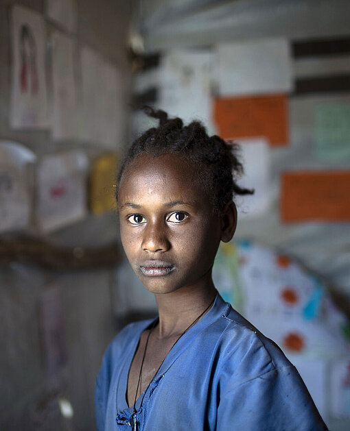 Eyerus lebt mit ihrer Großmutter in einem IDP-Camp in Shire. Ein Mädchen schaut ernst in die Kamera. Sie trägt ein blaues T-Shirt und ihre schwarzen Haare sind zu einem Zopf gebunden.