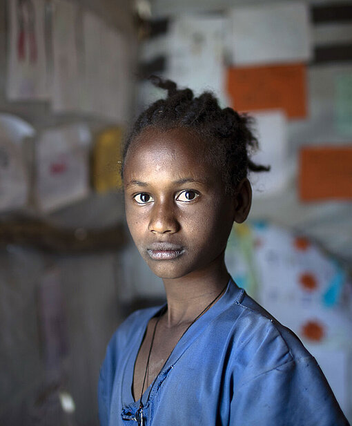 Ein Mädchen schaut ernst in die Kamera. Sie trägt ein blaues T-Shirt und ihre schwarzen Haare sind zu einem Zopf gebunden.