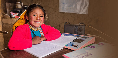 Ein Mädchen sitzt an einem Schreibtisch. Vor ihr liegt ein aufgeklapptes Heft, ein Ordner und ein Radio. Sie grinst in die Kamera und hält einen Stift in der Hand. 