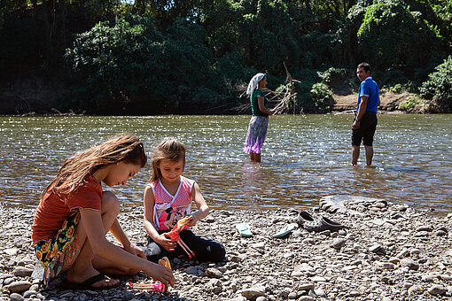 Zwei Erwachsene stehen im Fluss und angeln, zwei Mädchen sitzen am Ufer und spielen mit Puppen