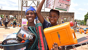 Zwei grinsende Frauen in Nigeria halten Hilfsgüter, darunter ein Kanister und ein Kochtopf.