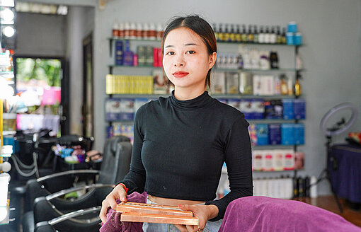 Eine Asiatin in schwarzem Pullover steht in einem Friseursalon und schaut zur Kamera