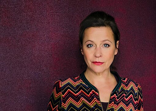 Jule Ronstedt Eine dunkelhaarige Frau mit einem bunt gemusterten Kleid steht in einer Bar