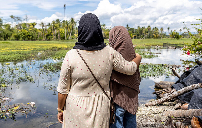 Farhana hat den Arm um die Schultern eines Mädchens gelegt. Sie schauen beide auf die überflutete Umgebung.