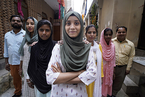 Die Mitglieder der Jugendgruppe aus Delhi stärken sich gegenseitig den Rücken Eine Gruppe junger Menschen steht zwischen Mehrparteienhäusern hintereinander. Sie zeigen ihre Solidarität füreinander, indem sie sich gegenseitig an die Schulter fassen. Ganz vorne steht selbstbewusst ein Mädchen mit verschränkten Armen