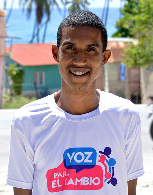 Carlos kam über einen Vortrag von Plan International zum Thema Kinderschutz und Aktivismus. Carlos steht draußen an einer Straße und lächelt in die Kamera. Er trägt ein weißes T-Shirt mit buntem Druck.