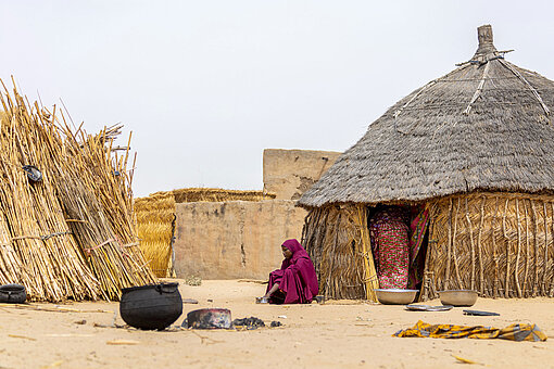 Zina lebt in einem abgelegenen Dorf in Niger Eine junge Frau in rotem Kleid und Kopftuch sitzt am Eingang einer Hütte. Im Hof stehen noch ein Kessel und große Schüsseln.