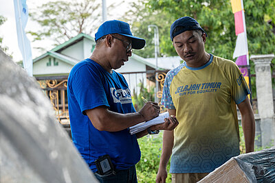 Zwei Männer stehen nebeneinander im Freien. Der eine trägt ein blaues T-Shirt und eine Kappe mit dem Schriftzug „Plan International“ und schreibt etwas in ein Notizbuch. Im Hintergrund sind ein Haus, Bäume und ein Zaun zu sehen. Die Szene vermittelt den Eindruck von Hilfsmaßnahmen oder Organisation vor Ort.