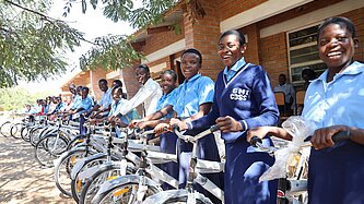 Eine Gruppe von Schülerinnen und Schülern, die lächelnd neben neu verteilten Fahrrädern vor einem Schulgebäude in Malawi stehen. Die Schülerinnen und Schüler tragen Schuluniformen und die Fahrräder sind bereit zur Nutzung.