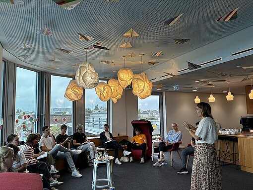 Mehrere junge Menschen sitzen in einem Raum, eine Frau führt einen Workshop durch