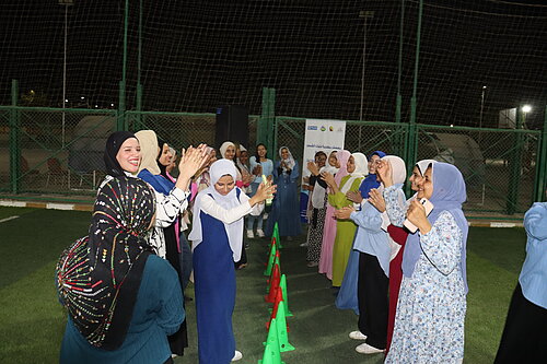 Eine Gruppe von Frauen und Mädchen steht in zwei Reihen auf einem Sportplatz. Sie lachen, klatschen begeistert und schauen sich gegenseitig an.