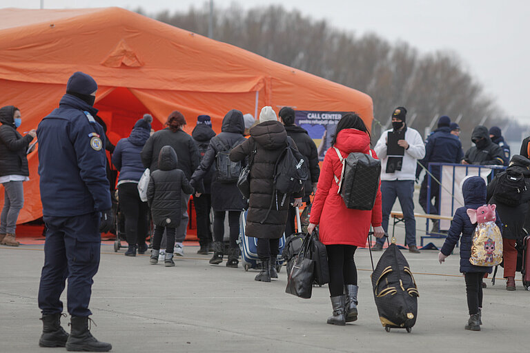 Menschen in dicken Jacken und mit Rucksack oder auch Koffern stellen sich vor einem orangenen Zelt in einer Reihe auf