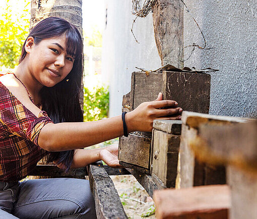 Eine junge Frau kniet vor Bienenstöcken und lächelt in die Kamera