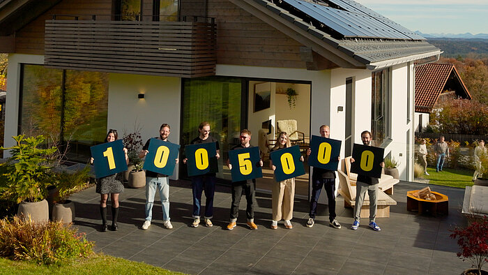 Eine Gruppe von Menschen hält große Schilder mit der Zahl "1005000" vor einem modernen Haus. Sie stehen in einer Reihe und präsentieren die erreichte Spendensumme der Omaze Traumhauslotterie für Plan. 