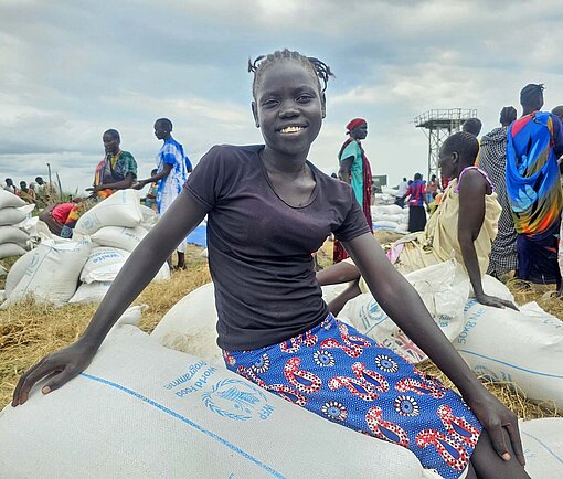 Damit sich die Menschen in Südsudan wieder selbst ernähren können, sind Projekte zur Gemeindeentwicklung erforderlich Eine Frau sitzt auf einem Sack Getreide und lacht erfreut