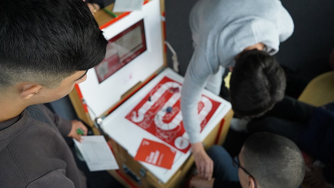 Drei Jungen beugen sich über die aufgeklappte Box, auf der ein rotes Brettspielfeld liegt.