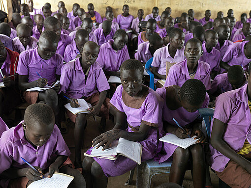 Seit einiger Zeit versucht er vor allem, seine Schüler:innen in der Schule zu behalten. Schüler:innen mit lilafarbener Uniform sitzen im Klassenzimmer und schreiben fleißig mit.