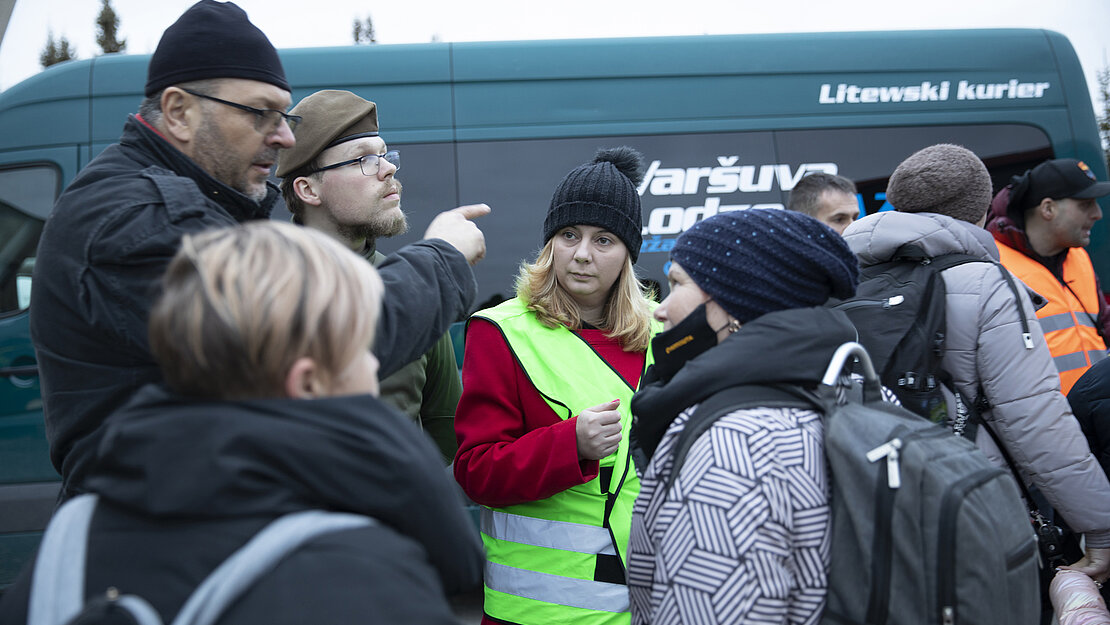 Monica koordiniert die Unterstützung für neu angekommene gefluechtete Menschen aus der Ukraine. Monica steht in eine gelbe Warnweste gekleidet vor einem Bus, inmitten von Menschen.