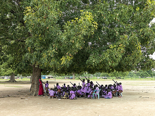 Wegen der Hungerkrise bleiben viele Kinder und Jugendliche dem Unterricht fern. Eine Schulklasse steht mit einer Lehrerin unter dem Baum. Viele Kinder haben ihren Arm gehoben und melden sich.