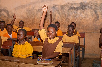 Mehrere Schüler:innen sitzen in einem Klassenraum in Ghana an Schultischen. Sie trragen alle eine Schuluniform, einige melden sich und lächeln.