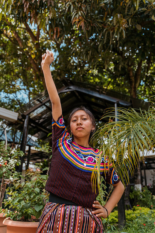 Eine junge Frau mit traditionell bestickter Kleidung steht zwischen Pflanzen. Sie hat eine Hand in die Hüfte gestemmt und streckt ihre Faust in die Luft
