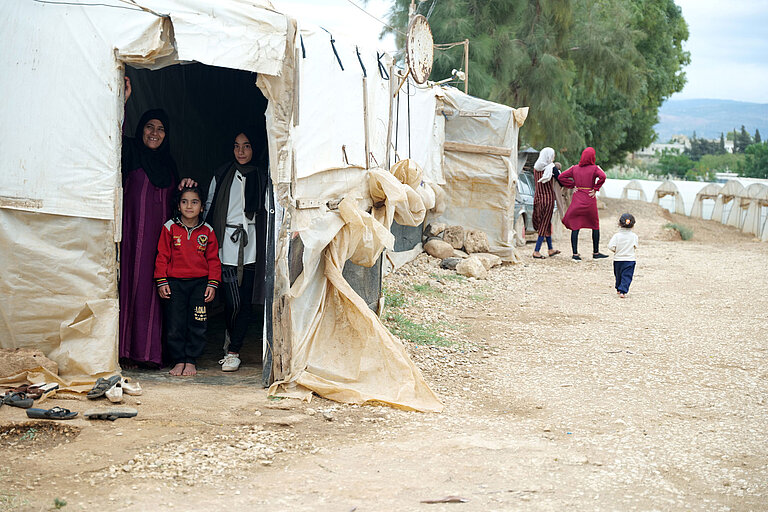 Im Vordergrund stehen zwei Frauen und ein Mädchen im Eingang eines Zeltes. Im Hintergrund sieht man den Rest des Zeltlagers, in dem auch Mädchen und Frauen herumlaufen.