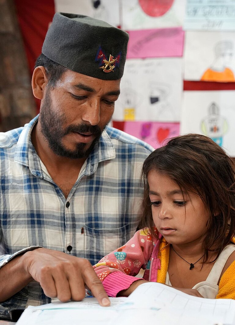 Vater sitzt neben seiner jüngsten Tochter und unterstützt sie beim Lesen eines Schulbuchs