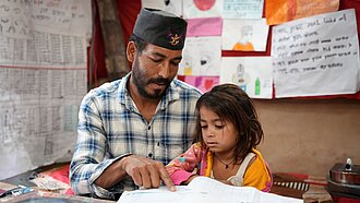 Vater sitzt neben seiner jüngsten Tochter und unterstützt sie beim Lesen eines Schulbuchs