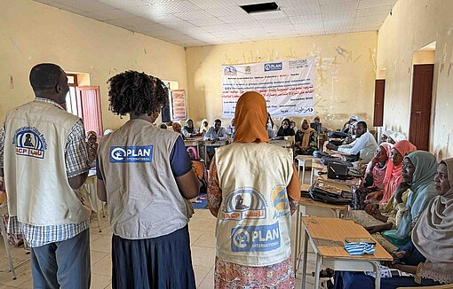 Drei Personen mit Plan-Logo auf der Kleidung stehen in einem Klassenraum und sprechen vor den dort sitzenden Erwachsenen, meistens Frauen mit Kopftüchern.