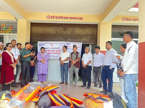 Eine Gruppe von Menschen, darunter Männer und Frauen, stehen vor einer Wand in einem Raum, in dem eine Veranstaltung zur Katastrophenvorsorge stattfindet. Sie hören einer Person zu, die spricht. Auf dem Boden liegen Rettungswesten.