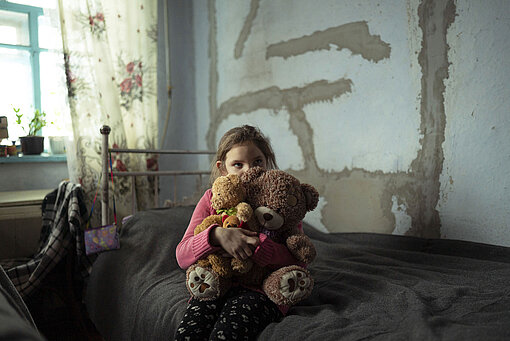 Natalka (9) kuschelt mit ihren Teddys auf ihrem Bett. Ein junges Mädchen sitzt auf einem Bett und hält zwei Teddys im Arm, an die sie ihr Gesicht schmiegt.