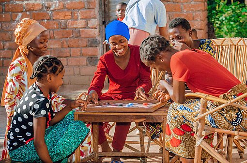 Junge Frauen in bunten Kleidern sitzen an einem Tisch und amüsieren sich bei einem Brettspiel