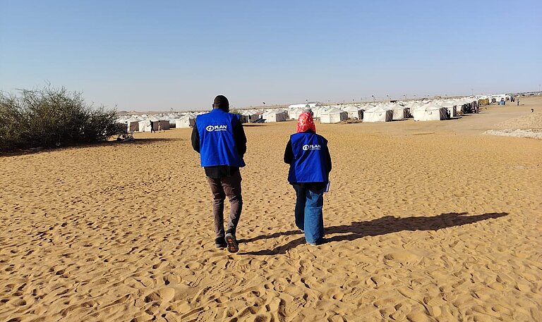 Zwei Mitarbeitende von Plan International laufen auf ein großes Geflüchtetencamp zu
