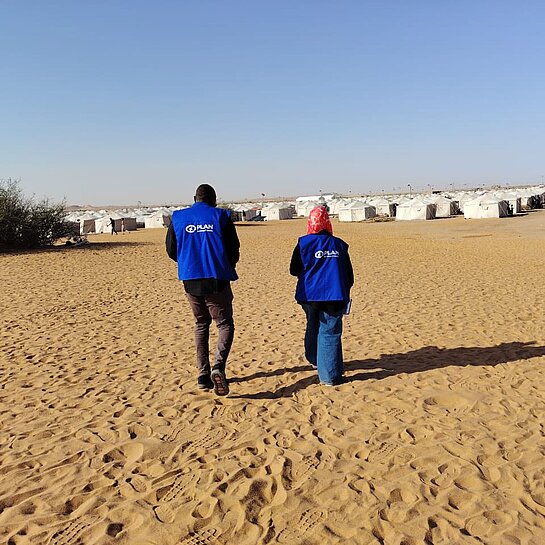 Zwei Mitarbeitende von Plan International laufen auf ein großes Geflüchtetencamp zu