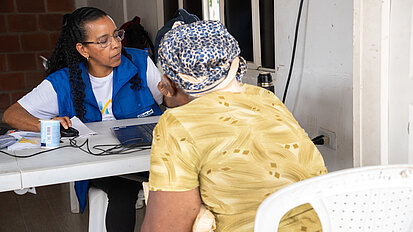 Eine Frau im blauen Plan International-Vest sitzt an einem Tisch und spricht mit einer älteren Frau mit Kopftuch, die ihr gegenüber sitzt. Auf dem Tisch liegen Dokumente und ein Laptop. Die Szene wirkt wie eine Beratung.