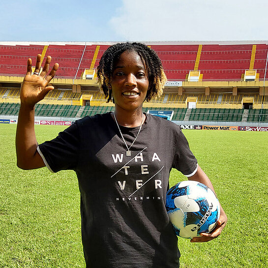 Eine junge Schwarze Frau steht mitten in einem Fußballstadion. Sie winkt in die Kamera und hat einen Ball im Arm.