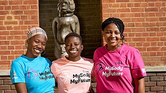 Drei junge Frauen aus Simbabwe halten sich in den Armen und lachen. Sie tragen T-Shirts mit der englischen Aufschrift "Mein Körper, meine Zukunft".