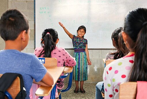 Ein Mädchen steht an der Tafel vor ihren Mitschülerinnen und Mitschülern und trägt etwas vor