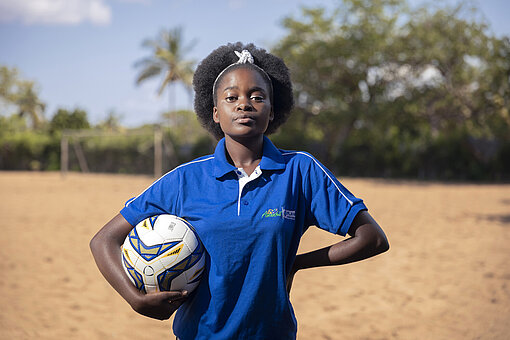 Eunice steht auf dem sandigen Fußballplatz, hat den Ball unter einen Arm geklemmt, die andere Hand in die Hüfte gestemmt und schaut in die Kamera