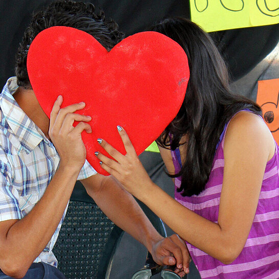 Ein Junge und ein Mädchen sitzen auf Stühlen und stecken die Köpfe zusammen, vor ihre Gesichter halten sie ein Herz aus Pappe.