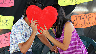 Ein Junge und ein Mädchen sitzen auf Stühlen und stecken die Köpfe zusammen, vor ihre Gesichter halten sie ein Herz aus Pappe.